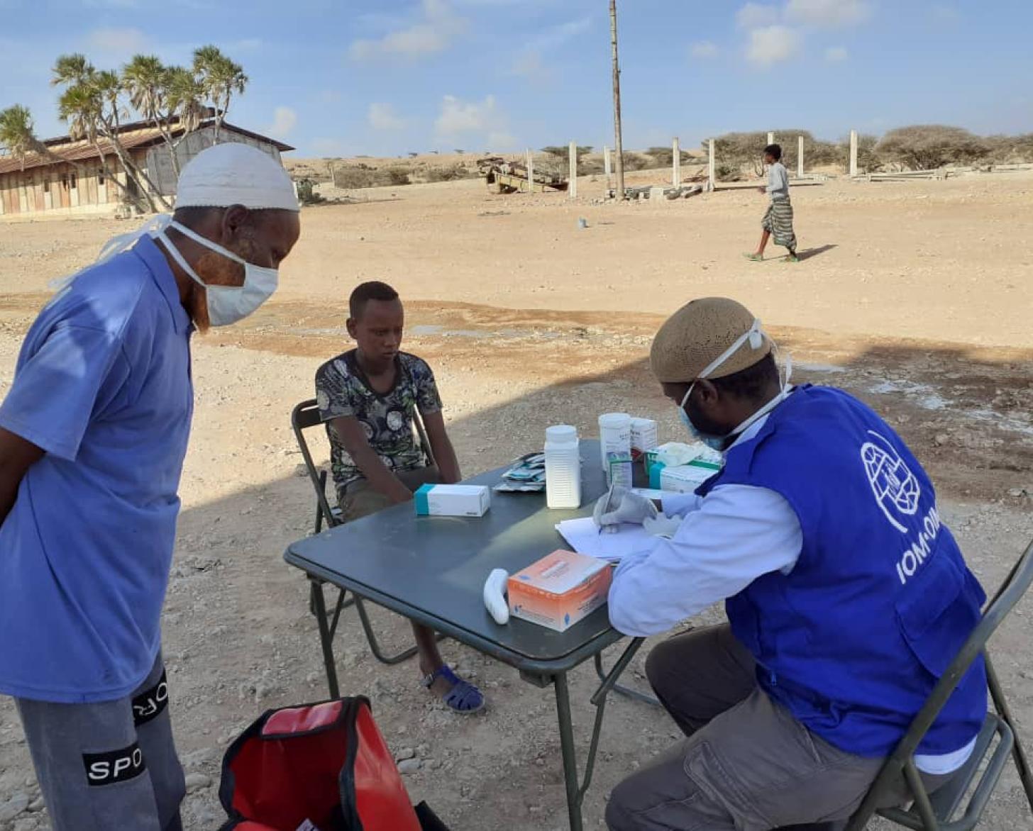 Un miembro del personal de la ONU y una mujer están sentados uno frente al otro, en una mesa que hace las veces de escritorio; mientras el funcionario toma notas y otro hombre con una mascarilla protectora observa el proceso.