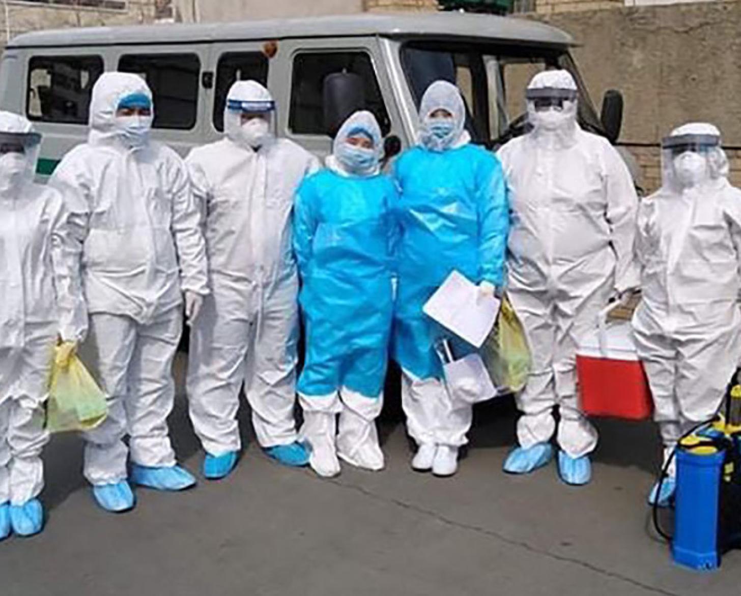 El equipo del Centro Nacional de Enfermedades Transmisibles se encuentra de pie, uno al lado del otro, delante de una furgoneta con un equipo de protección completo de pies a cabeza.