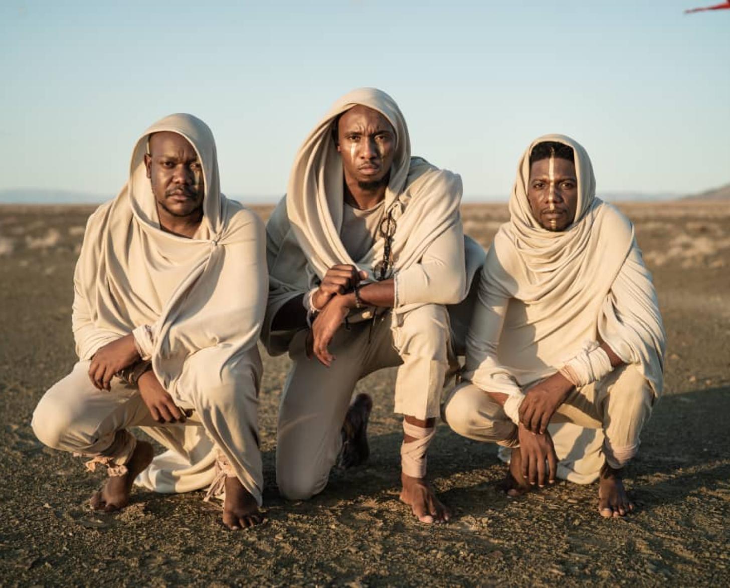 The trio band are shown crouched on the ground on what looks like an open field.