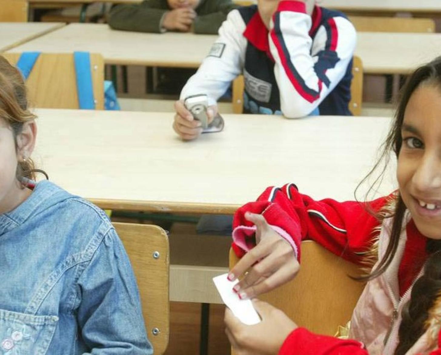 Roma children pose and smile at the camera, while sitting in their classroom.