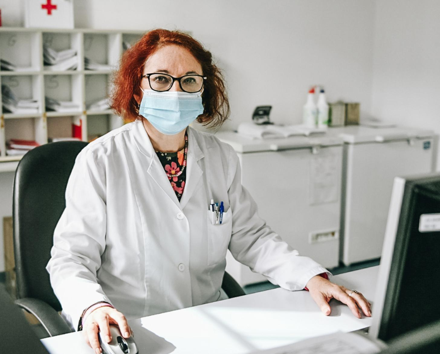 Snežana Bursać Aranđelović está sentada en su escritorio con una computadora (frente a ella) en el Instituto de Salud Pública del Cantón de Sarajevo. Lleva una bata blanca y una mascarilla quirúrgica azul y está mirando a la cámara. Podemos ver detrás archivos y mobiliario de oficina colocado en estanterías blancas.