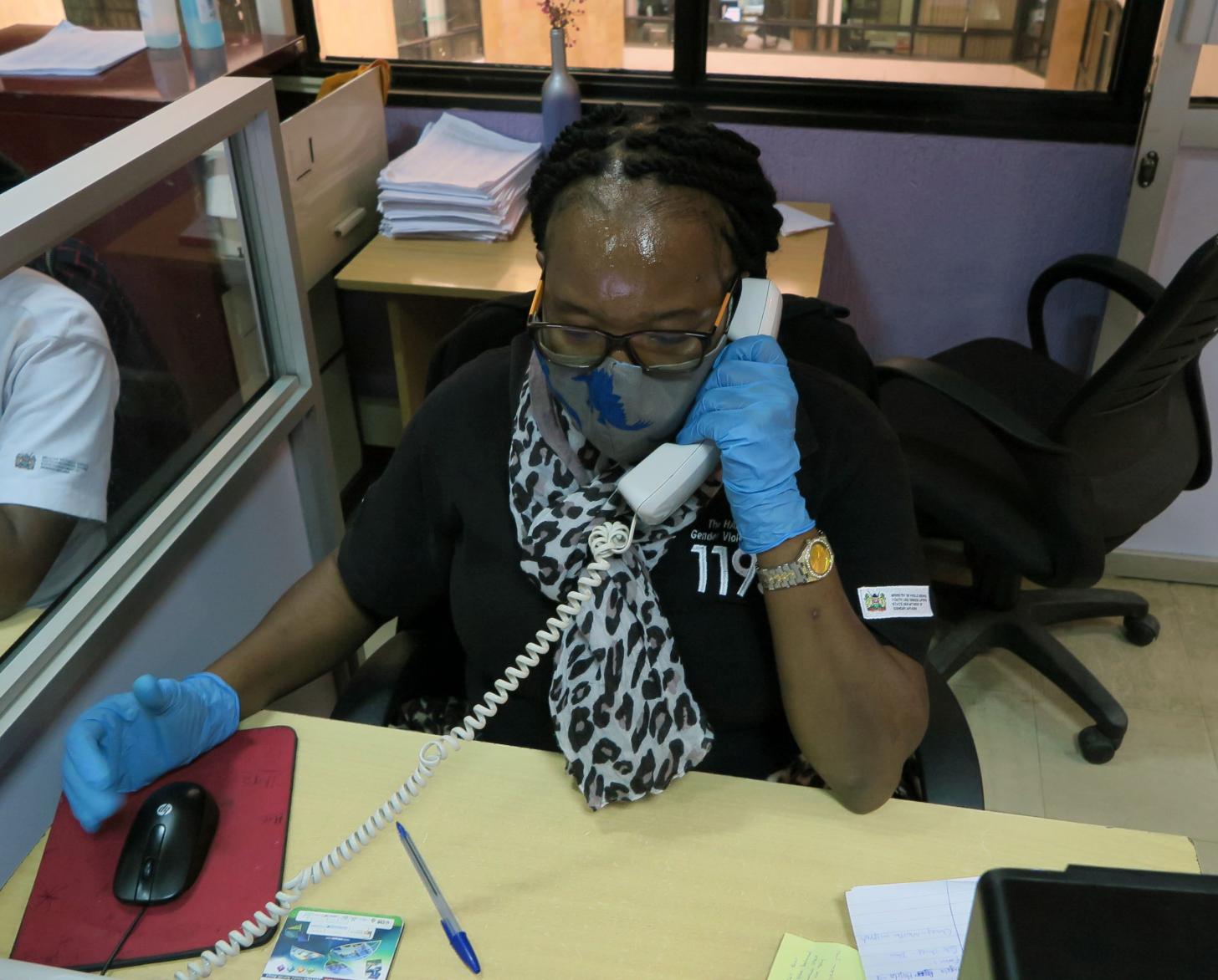 Una representante del centro de llamadas responde a una llamada realizada a la plataforma telefónica del servicio de atención a víctimas. Ella se sienta en un escritorio y usa una mascarilla y guantes protectores.