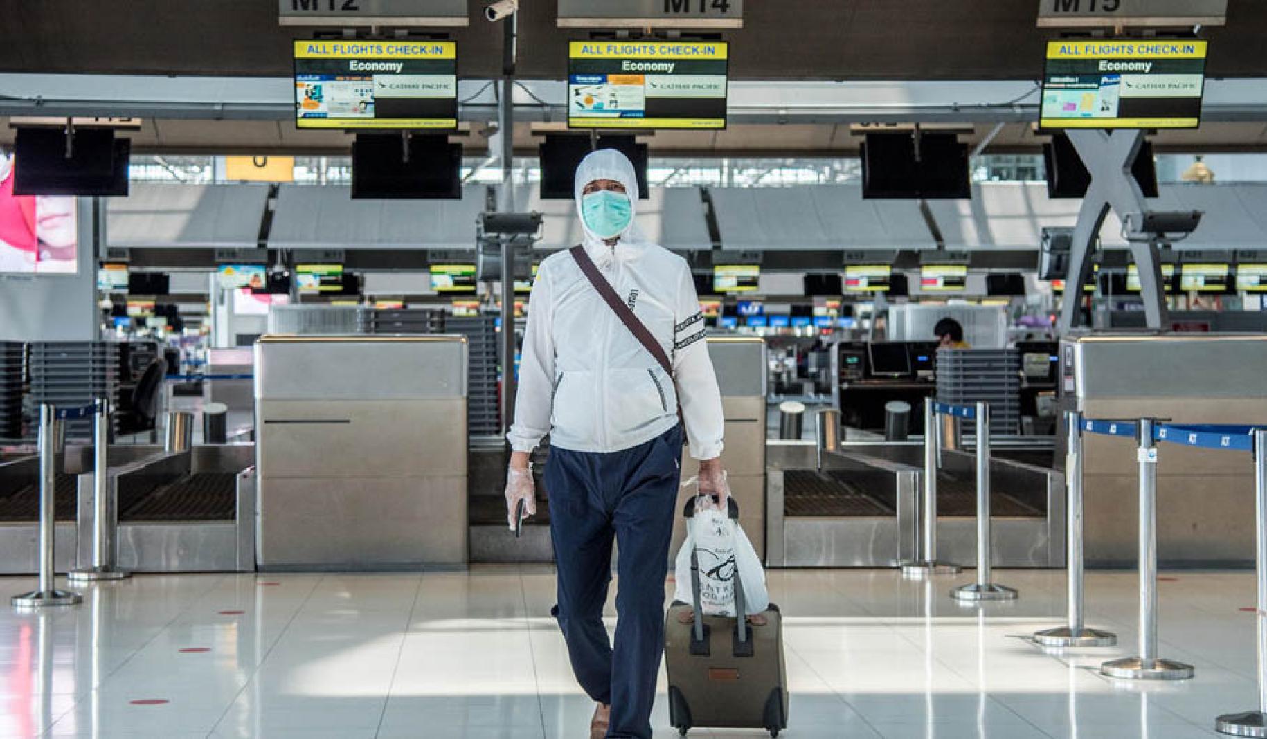 Un homme vêtu d'un survêtement bleu et blanc et portant un masque de protection respiratoire et des gants de protection transparents tire sa valise à roulettes devant un comptoir d'enregistrement de l'aéroport Suvarnabhumi de Bangkok, en Thaïlande.