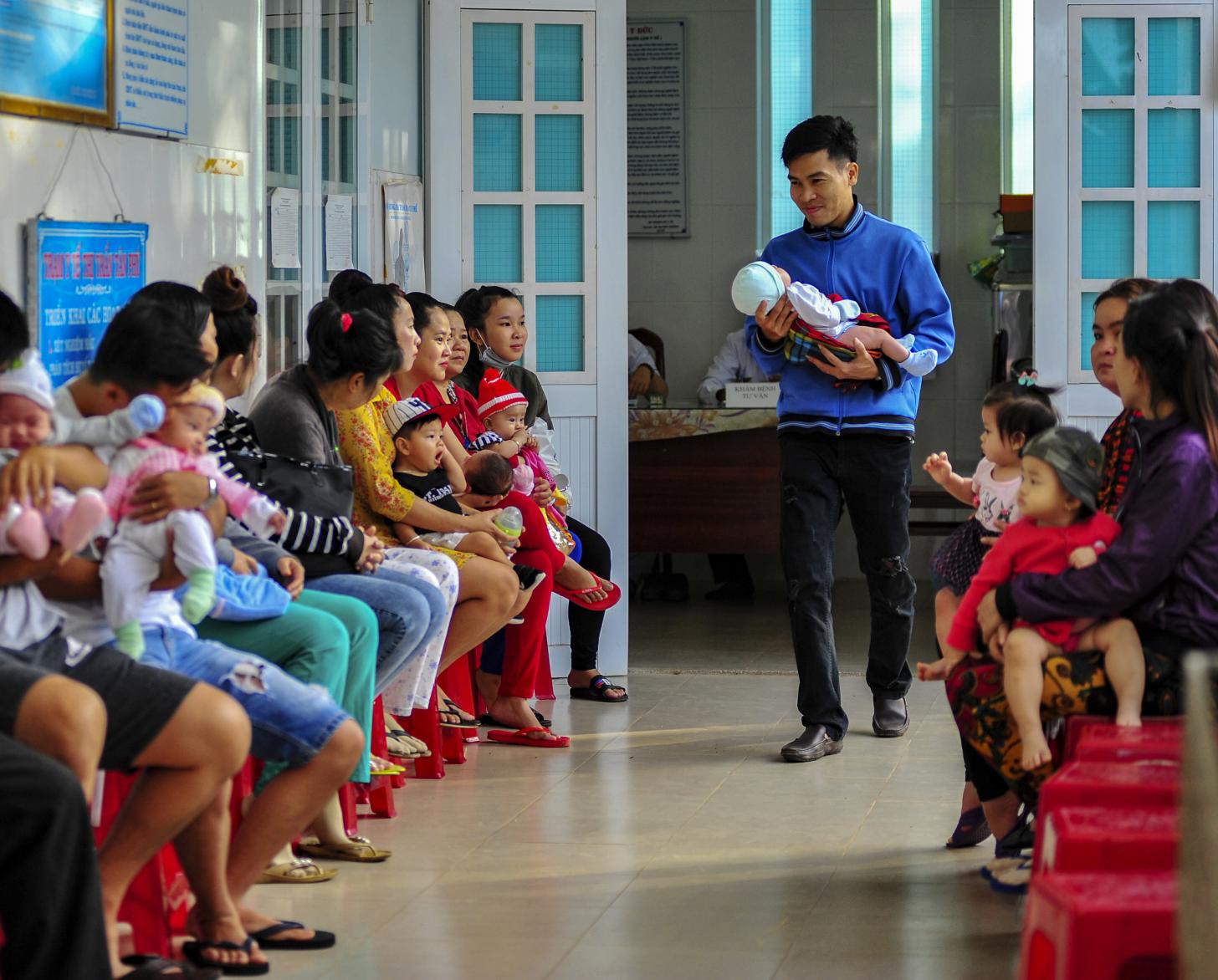 A father holds his newborn baby in the waiting space of a vaccination clinic where other parents sit along the hallway with their babies.