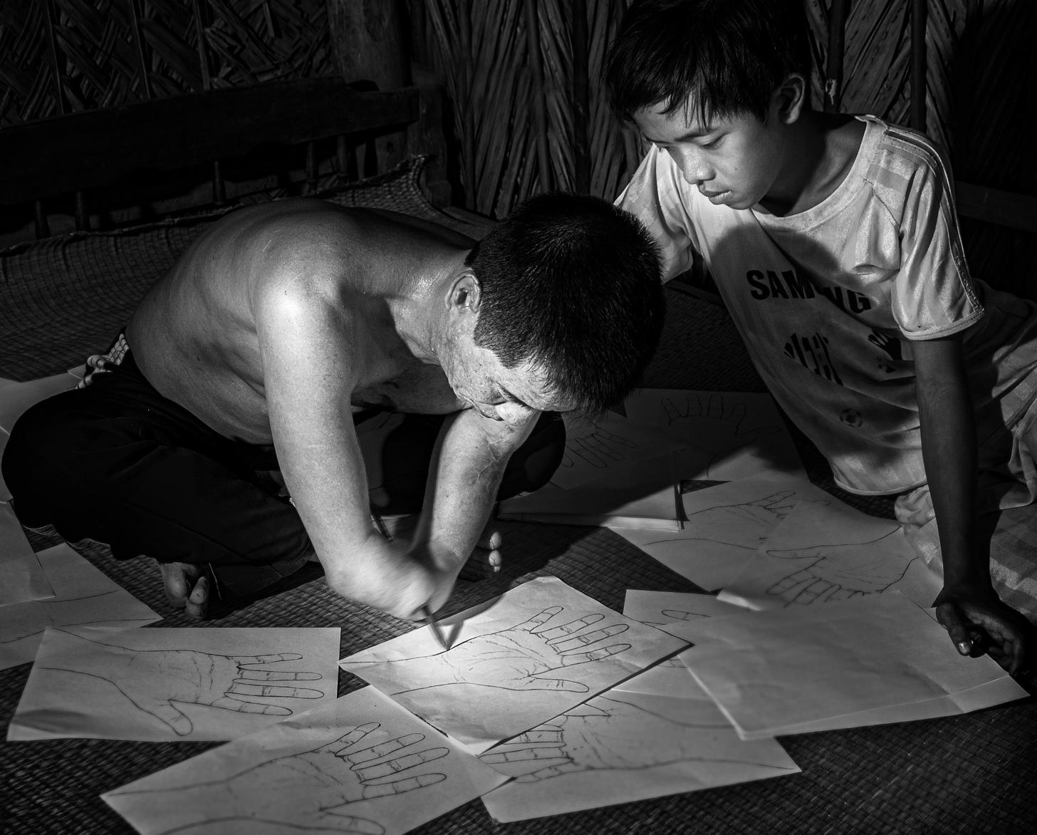 Black and white photo of a man and boy. The man has an amputated hand, sit together on the ground drawing and writing on sheets of paper.