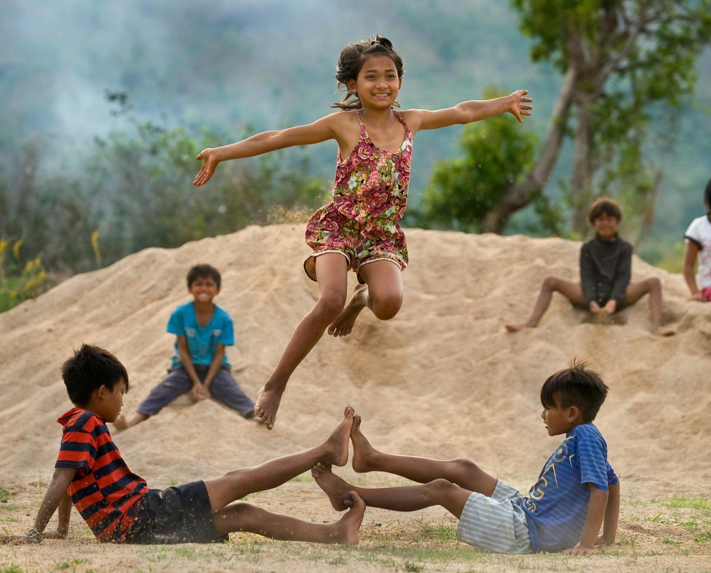 Children play outside. Two boys are on the ground touching their feet, while one girl is frozen in the air happily jumping above them.
