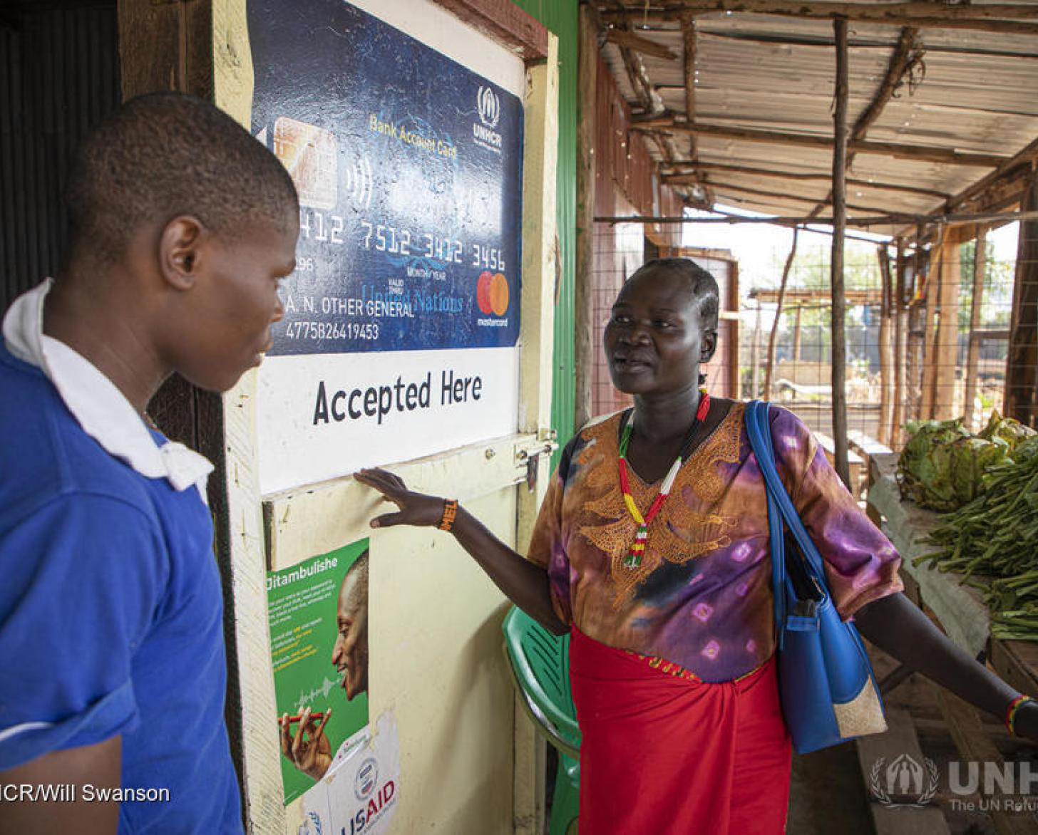 A woman stands by a walk covered in signage while speaking with a man, which stands by the doorway of the market at the Kalobeyei Settlement.