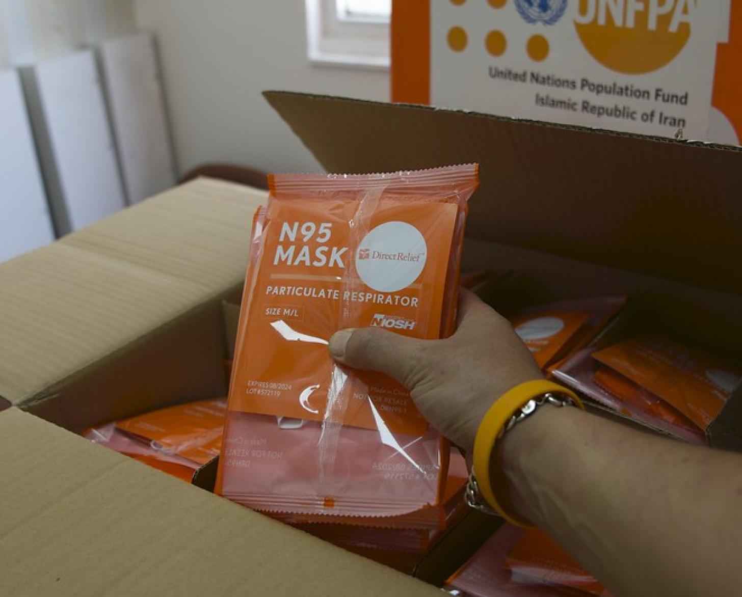 A hand holds up packaged PPE above a UNFPA-donated box.