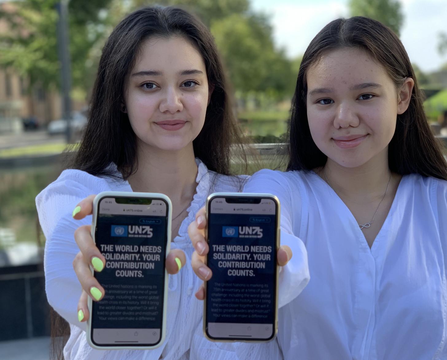 Two adolescent girls hold up their mobile phones directly towards the camera as they show the UN75 survey on the screen.