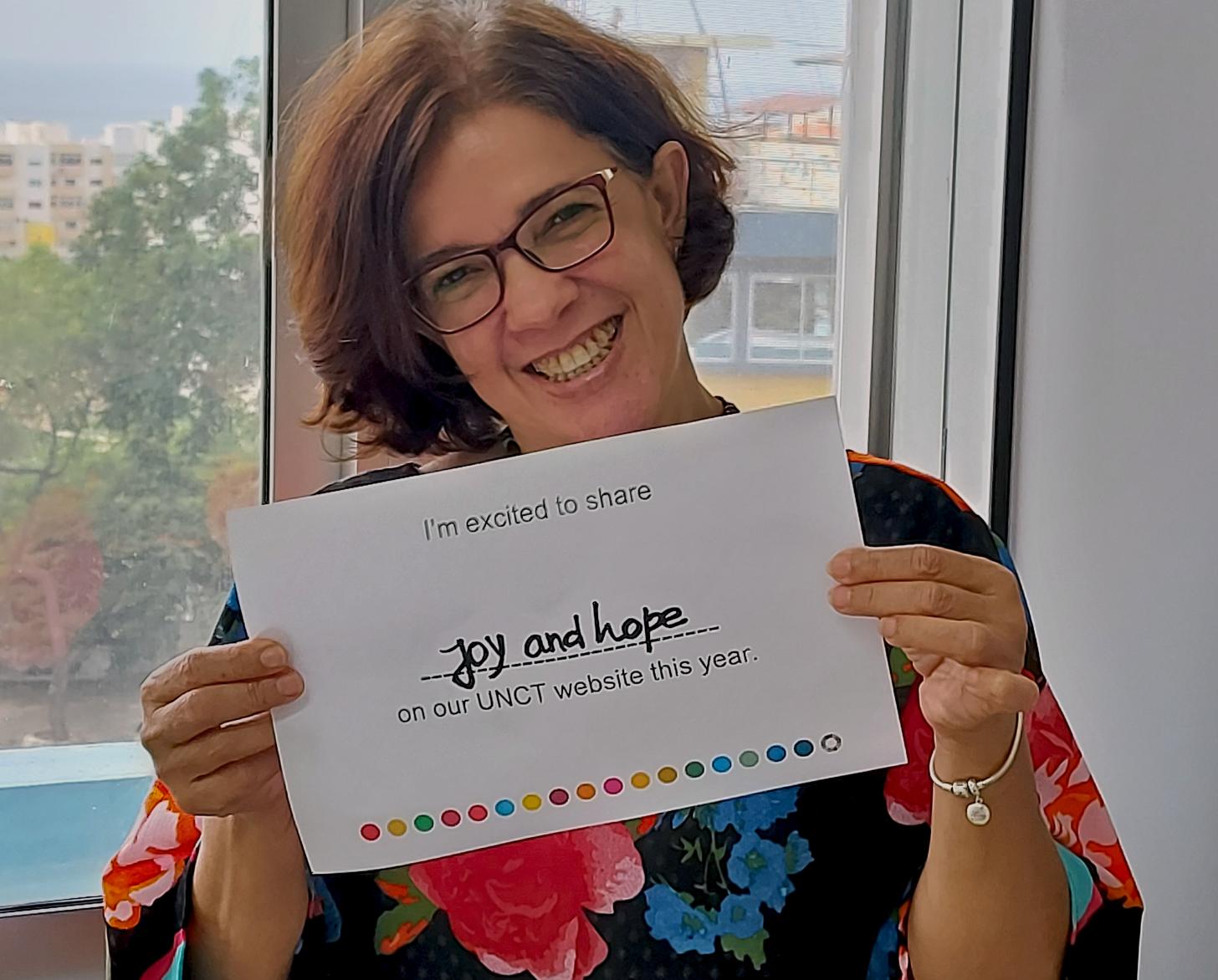 Anita warmly smiles as she holds a sign that reads she's excited to share joy and hope on their country team website.