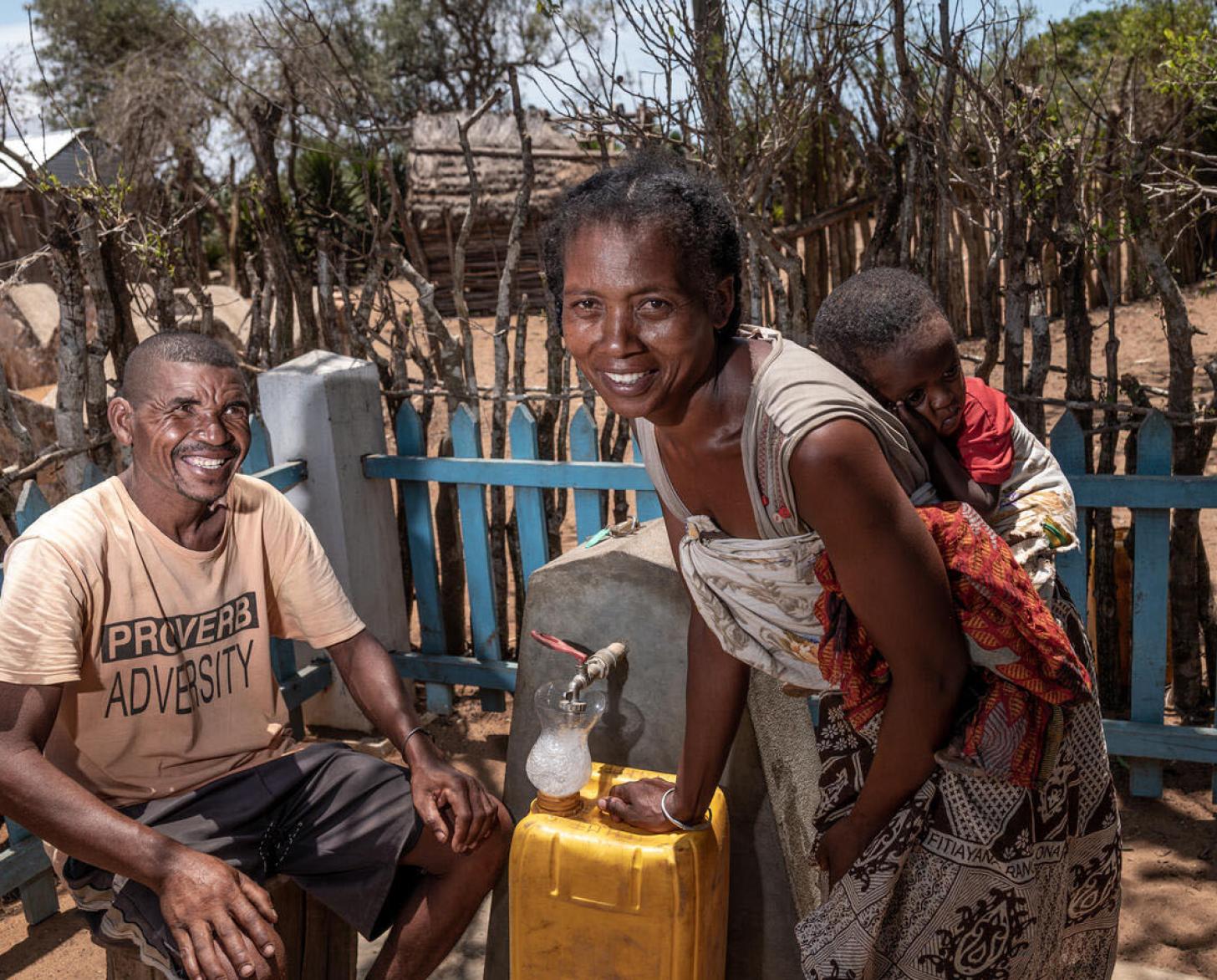 Una mujer que lleva a su bebé dormido a la espalda, llena un bidón de agua, y un hombre sentado a su lado, sonríen alegremente a la cámara. Ambos se encuentran junto a un grifo comunitario de agua.