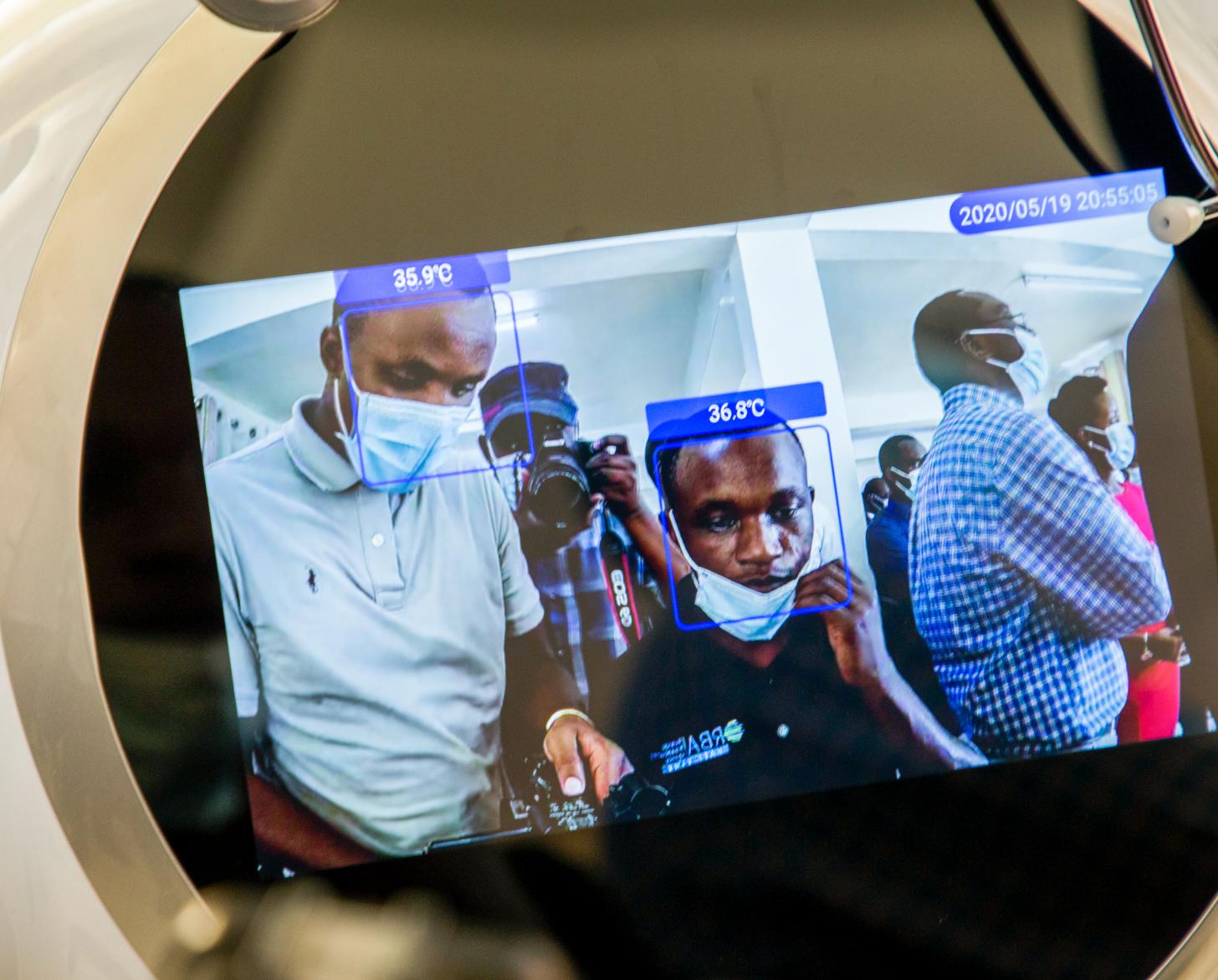 The screen of one of the robots shows the faces of two gentlemen with their body temperatures noted on the screens above their heads.