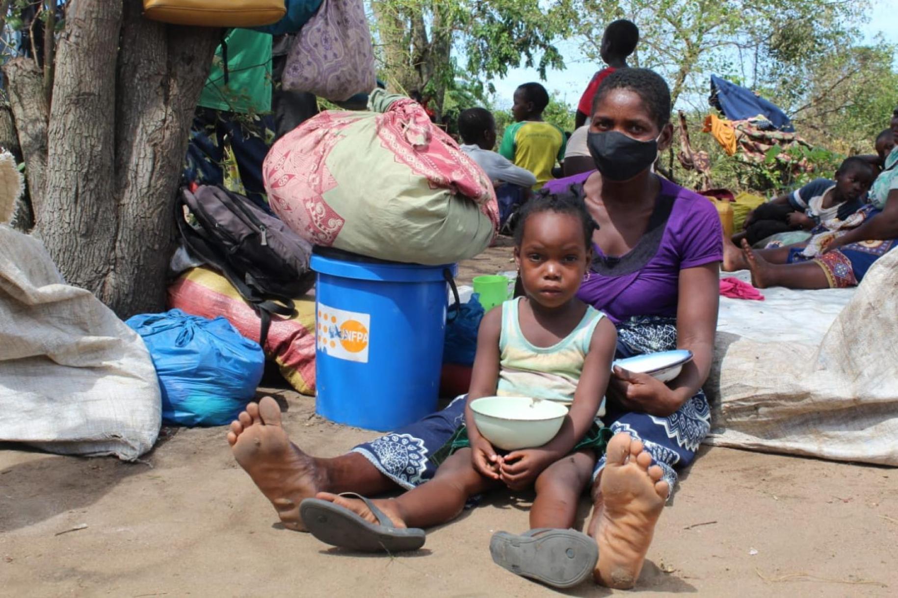 Una madre y un niño se sientan juntos en el suelo sosteniendo cuencos para la comida con las pertenencias a la izquierda de ellos, incluido un kit de dignidad del UNFPA.