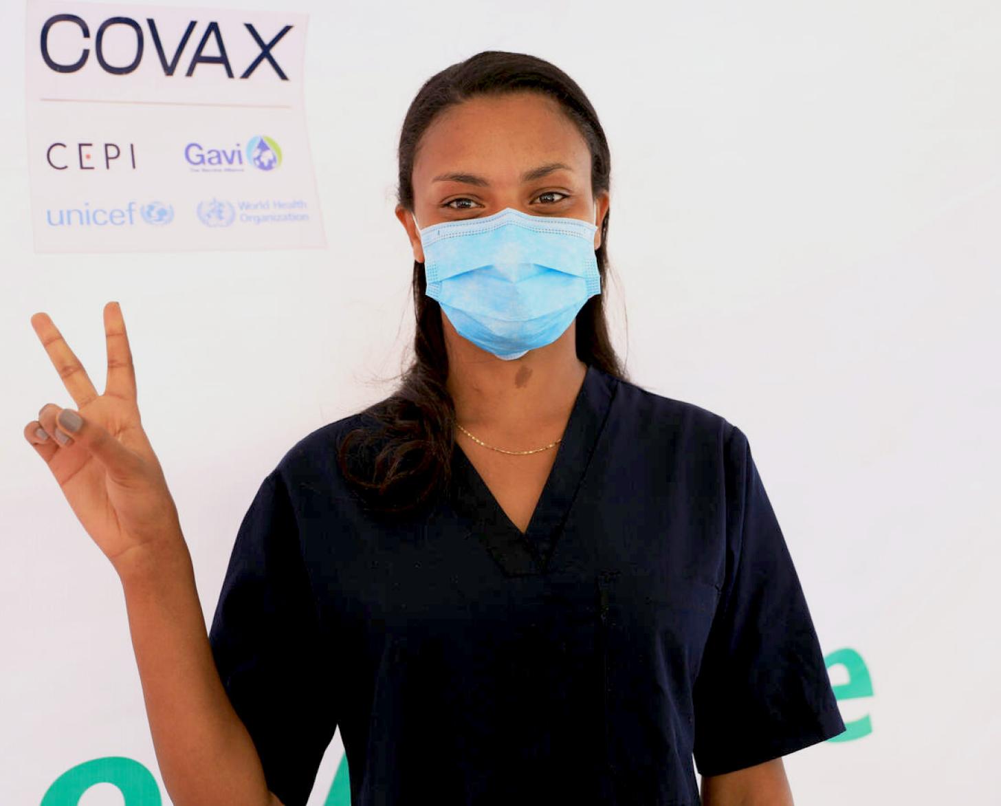 A healthcare professional stands by a COVAX sign and flashes a 'v' with her fingers to signify she was vaccinated.