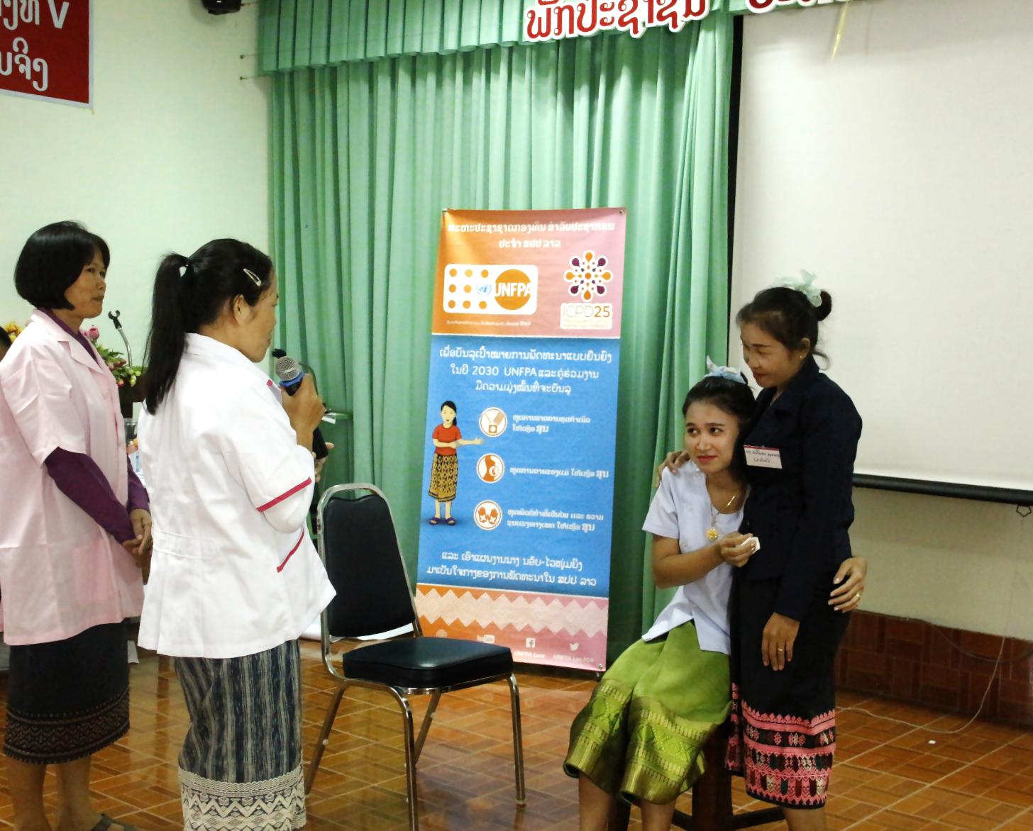Two medical professionals, in lab coats, speak to two young women hugging each other. 