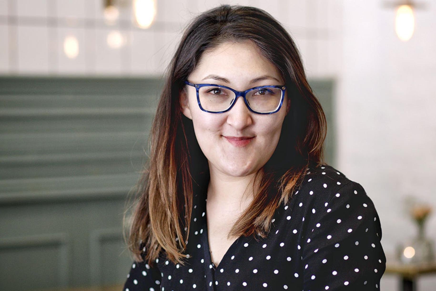 Una mujer con cabello castaño y gafas sonríe a la cámara con una camisa negra de lunares.