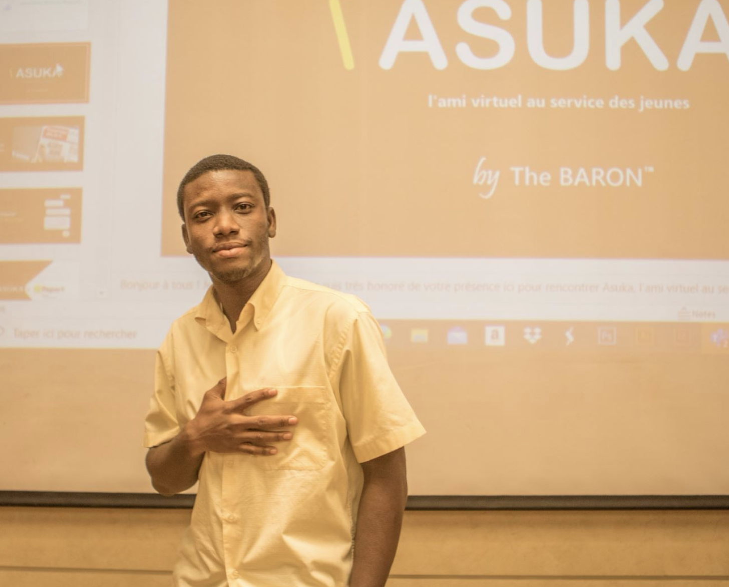 A young man, in a white button down shirt, stands in front of a projector holdings his hand to his heart. 