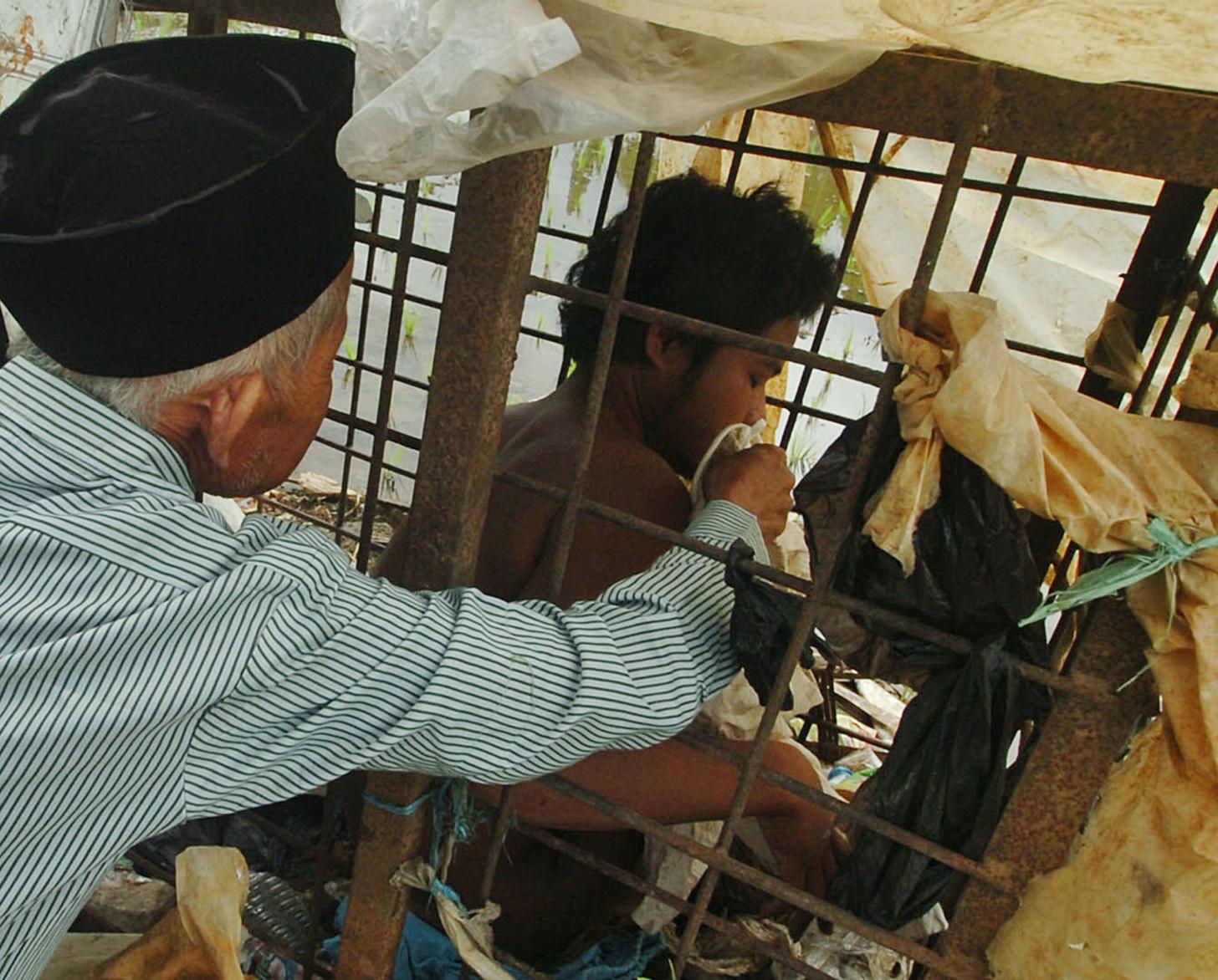 A young man in a cage is being helped by an older man in a black hat outside of the cage. 