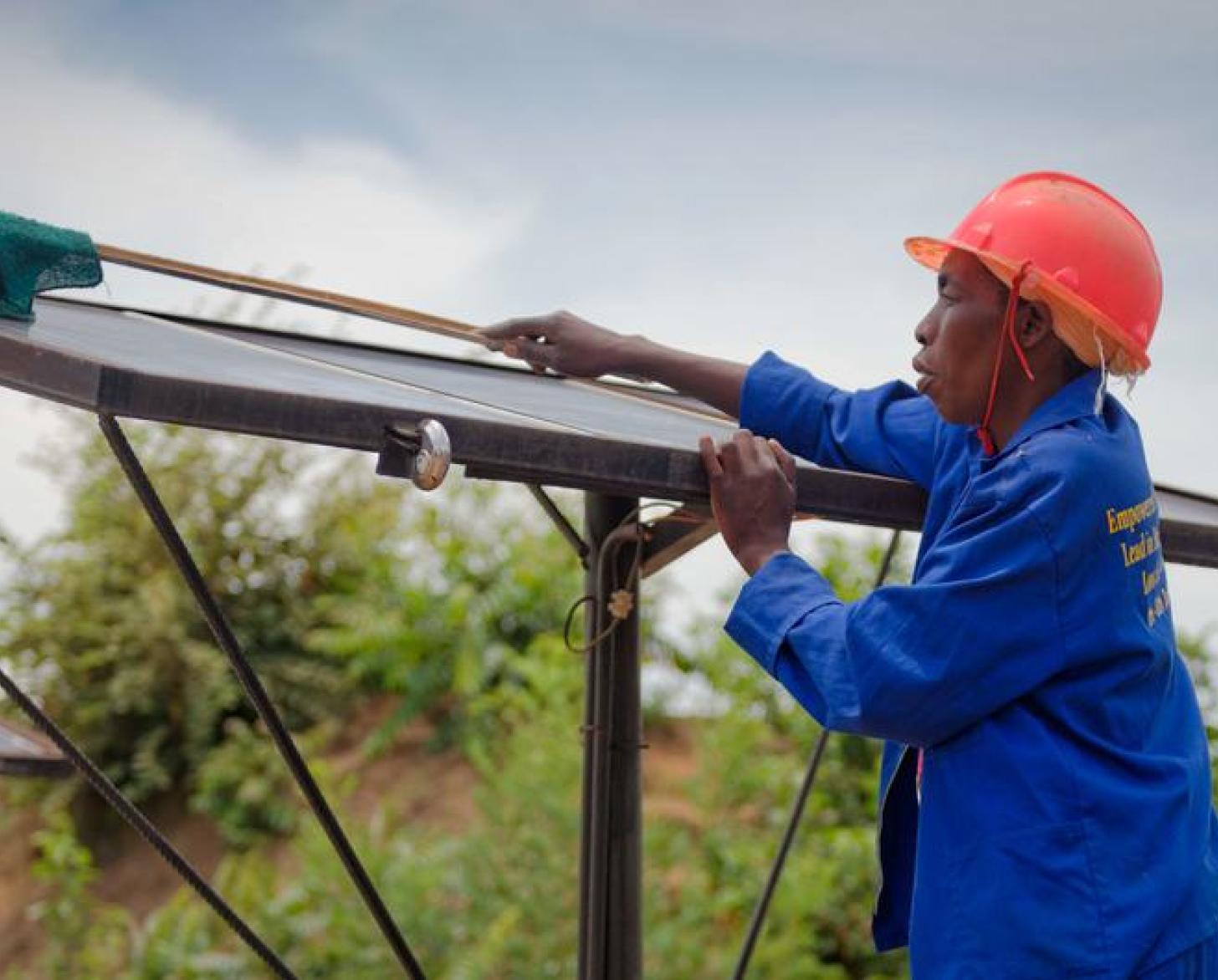 Un homme portant un bleu de travail et un casque de protection rouge nettoie un panneau solaire en extérieur.