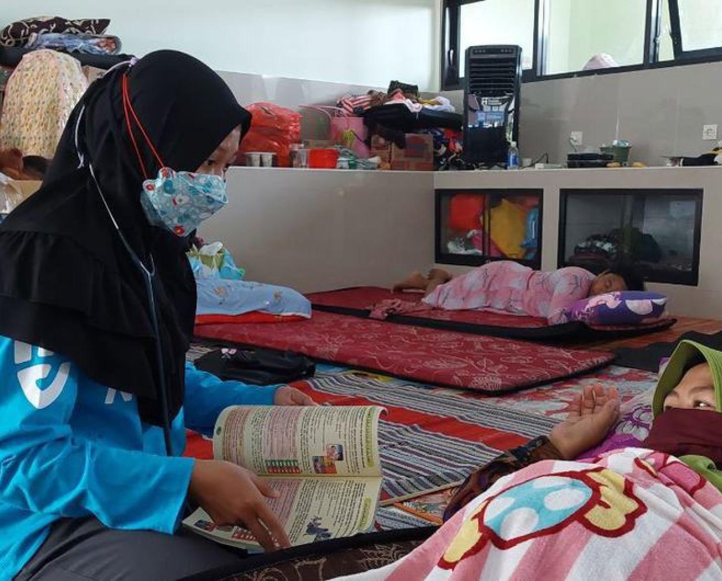 Une femme portant un voile noir sur la tête et un masque de protection respiratoire est assise en tailleur, à même le sol, et prend soin d'une femme enceinte allongée au sol dans une pièce entièrement tapissée.