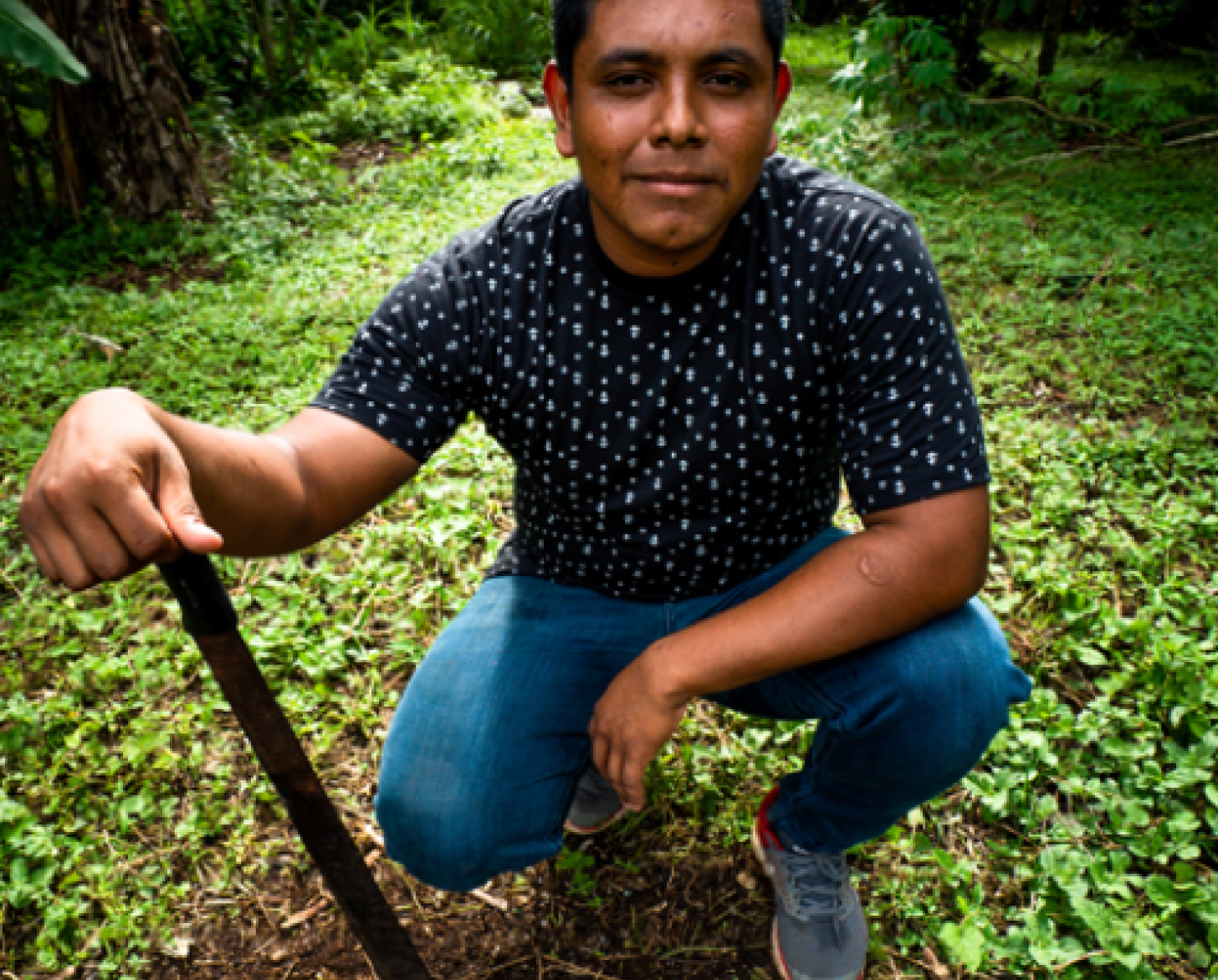 Un joven se agacha cerca de una pala en un terreno verde con árboles.