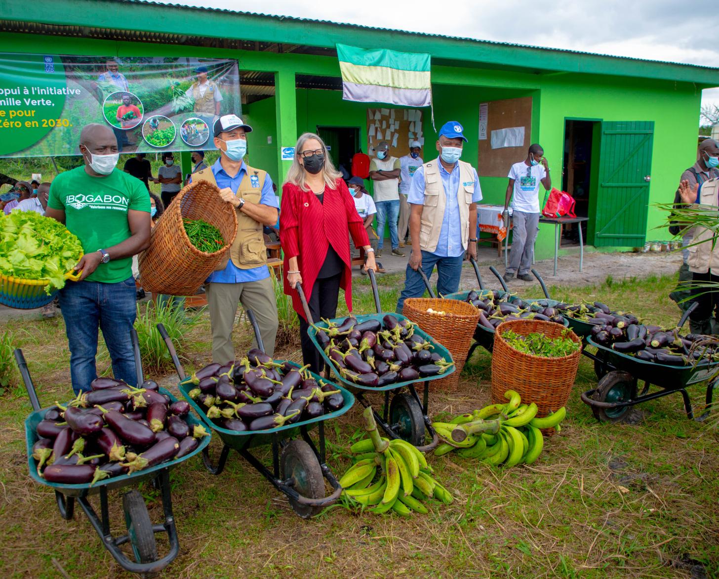 Plusieurs personnes portant un masque de protection respiratoire, dont la Coordonnatrice résidente de l’ONU au Gabon, Savina Ammassar, posent devant la caméra avec des charrettes et des paniers pleins de légumes et de fruits frais devant un bâtiment sur la façade duquel est apposé le drapeau du Gabon ainsi qu’une banderole indiquant "Projet d’appui à l’initiative Gabon famille verte".