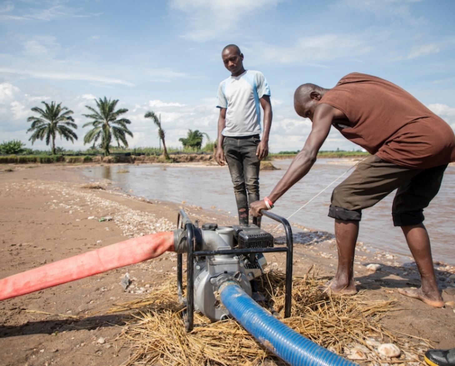 Au Burundi, au bord d'un étang bordé de palmiers, un homme se penche sur le moteur d'un système de collecte d'eau pour le faire fonctionner, sous le regard d'un autre homme.