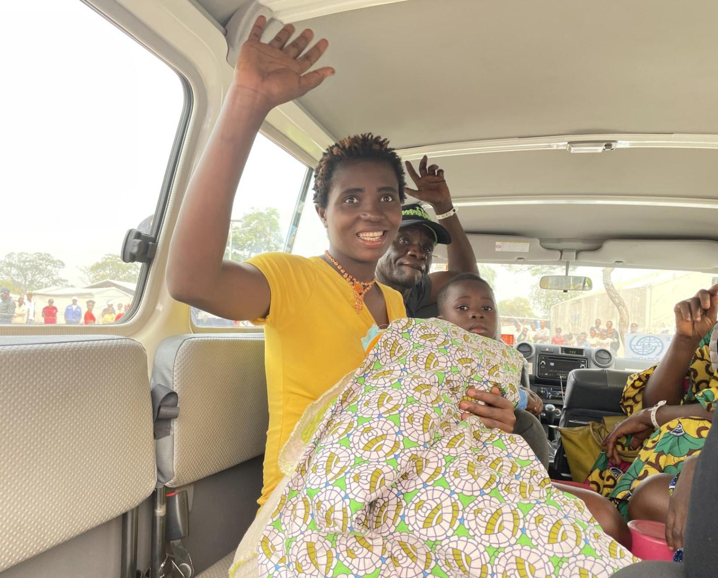 En Angola, une femme congolaise aux cheveux courts et vêtue d’un haut de couleur jaune à manches courtes est assise à bord d’un véhicule avec d’autres personnes, un bébé emmitouflé dans une couverture sur les genoux, et fait un signe d’adieu de la main droite en souriant.