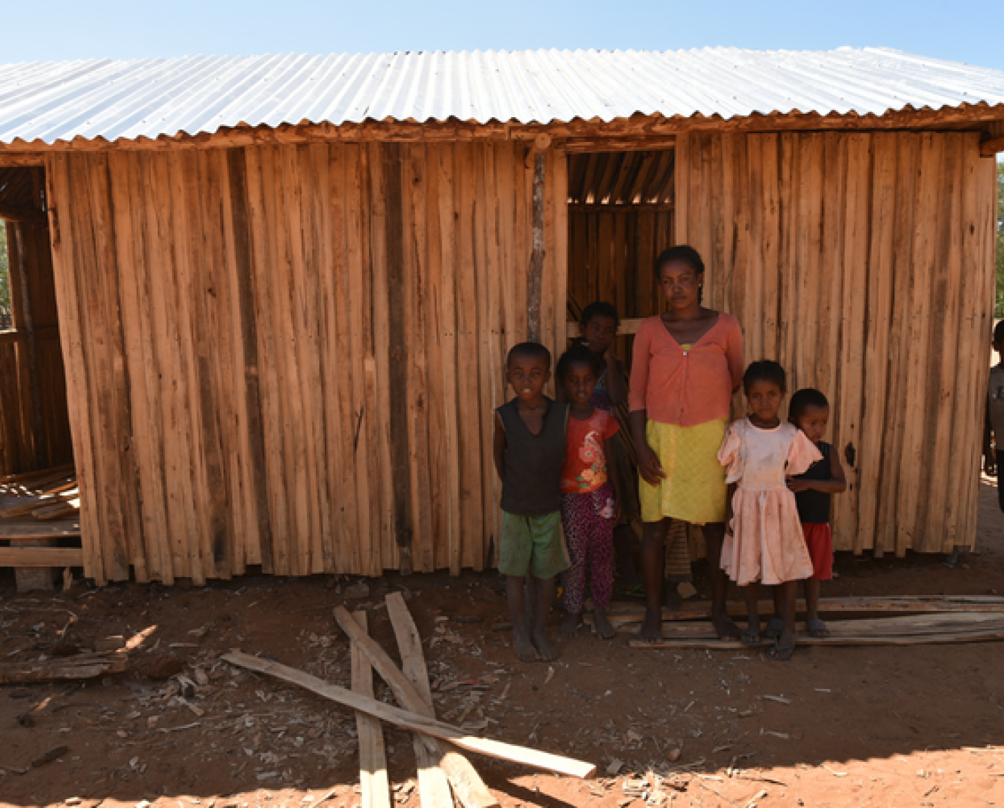 Une femme malgache se tient debout, devant la caméra, entourée de jeunes enfants, devant une modeste maison en bois, dans le village d’Amhamahavel, à Madagacar.