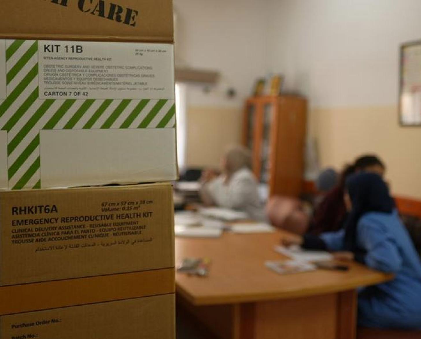 UNFPA kits and boxes, with women, not in focus, seated and talking in the background. 