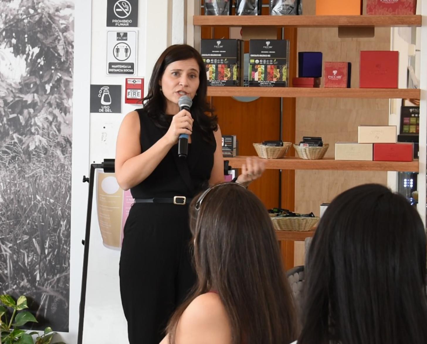 A woman in a black dress speaks into a microphone while other women listen to her