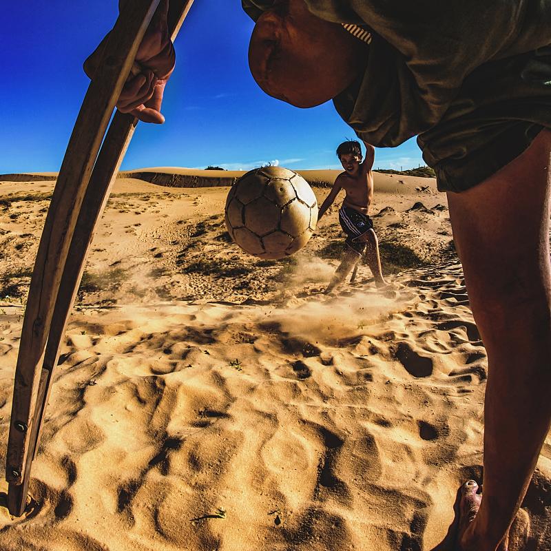 Photo shows a close-up between the legs of an man with an amputated leg. Through this angle you can see a soccer ball being kicked towards the man by a young boy.