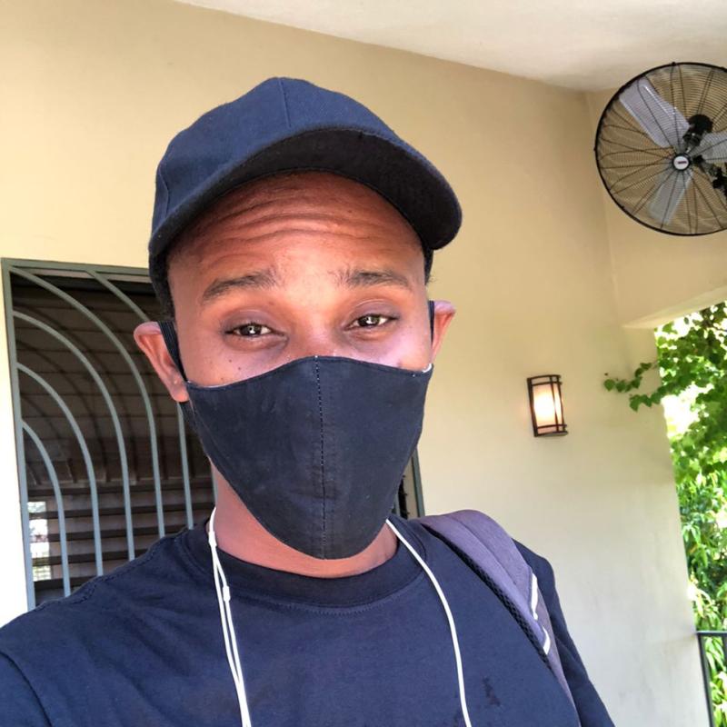 Un jeune homme portant un masque de protection respiratoire noir, une casquette de baseball noire et une chemise noire regarde droit dans l'objectif.